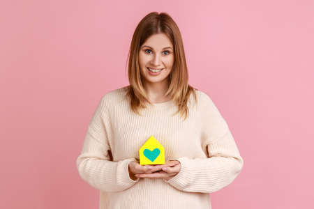 Portrait of positive friendly blond woman holding paper house with painted heart and looking at camera, charity, shelter, wearing white sweater. Indoor studio shot isolated on pink background.の写真素材