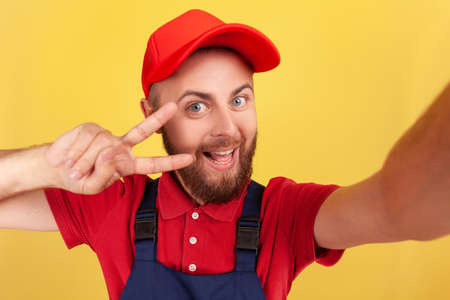 Smiling positive man courier or repairman broadcasting livestream or making point of view photo, showing v sign, wearing blue overalls and red cap. Indoor studio shot isolated on yellow background.の写真素材