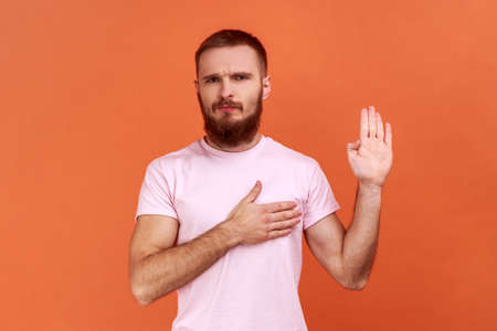 Portrait of serious young bearded man holding on chest another, swearing in honesty and devotion, patriotism, wearing pink T-shirt. Indoor studio shot isolated on orange background.の写真素材