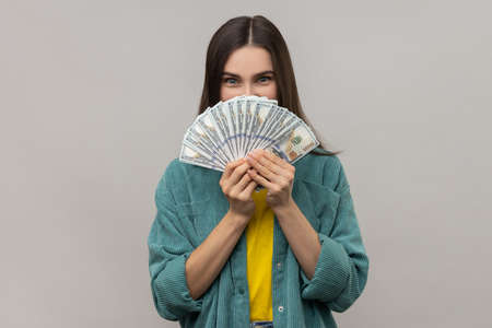 Portrait of attractive young adult woman covering half of face with many dollar banknotes, looking at camera, wearing casual style jacket. Indoor studio shot isolated on gray background.の写真素材