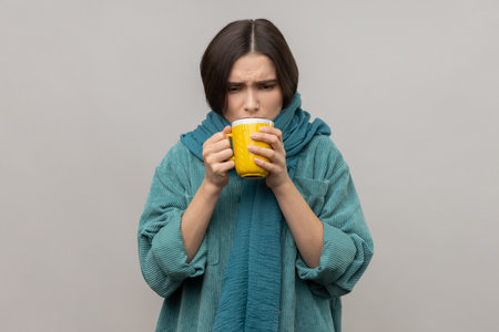 Sick unhealthy woman in scarf holding in hands cup with hot tea, feeling chills, drinking hot beverage, wearing casual style jacket. Indoor studio shot isolated on gray background.の写真素材