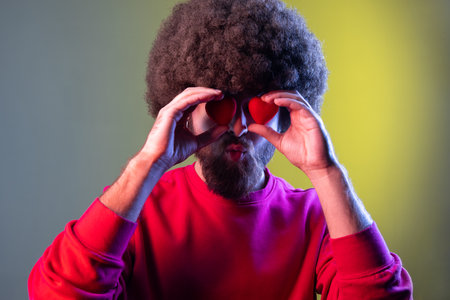 Portrait of hipster man with Afro hairstyle covering eyes with small red hearts, expressing romantic...