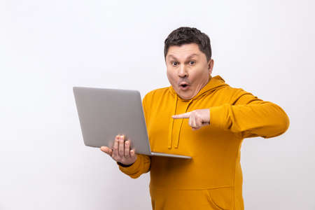 Astonished man working on laptop typing email or chatting in social network, pointing at display, shocked face expression, wearing urban style hoodie. Indoor studio shot isolated on white background.の写真素材