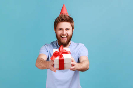 Portrait of man in party cone giving birthday gift and smiling, sharing present, donating and offering surprise in box, congratulation on anniversary. Indoor studio shot isolated on blue background.の写真素材