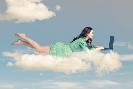 Hovering in air. Happy cheerful girl ruffle dress levitating with laptop, typing keyboard, reading good news message on computer while flying in mid-air. collage composition on day cloudy blue skyの写真素材