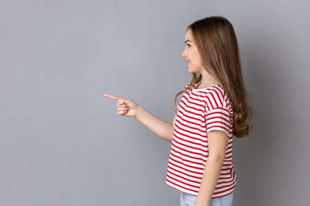 Side view portrait of little girl wearing striped T-shirt standing pointing aside presenting copy space for advertisement or promotional text. Indoor studio shot isolated on gray background.の写真素材