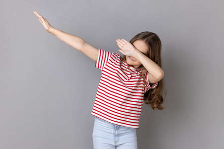 Winning, success gesture. Portrait of little girl wearing striped T-shirt showing dab dance pose,...