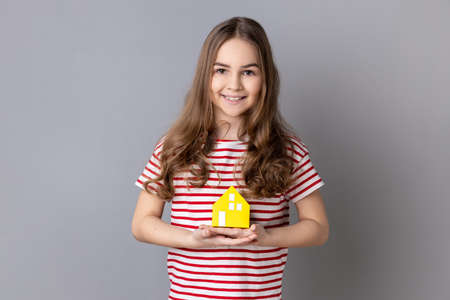 Portrait of charming little girl wearing striped T-shirt holding paper house and smiling to camera, advertisement of trustworthy real estate agency. Indoor studio shot isolated on gray background.の写真素材