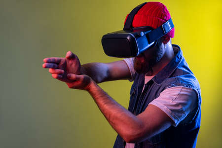 Hipster man with virtual reality glasses playing shooter game, pointing finger gun, killing in cyberspace using VR headset technology. Indoor studio shot isolated on colorful neon light background.の写真素材