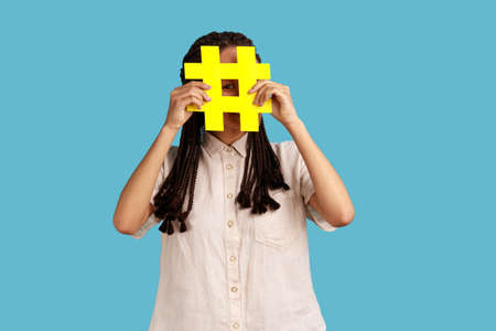 Funny woman blogger or smm manager with dreadlocks hiding face behind yellow hashtag sign, teaching to tag topics on websites, giving recommendations. Indoor studio shot isolated on blue background.の写真素材