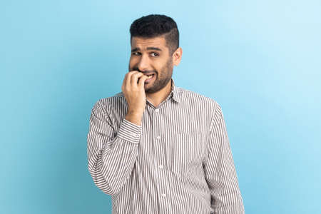 Portrait of stressed out worried bearded businessman biting nails, nervous about troubles, panicking and looking scared, wearing striped shirt. Indoor studio shot isolated on blue background.の写真素材