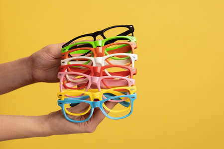 Profile side view closeup of woman hand holding and showing many colorful different eyeglasses. Indoor studio shot isolated on yellow background.の写真素材