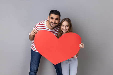 Portrait of optimistic positive father and daughter in striped T-shirts holding big red hear, looking at camera, expressing love to each other. Indoor studio shot isolated on gray background.の写真素材