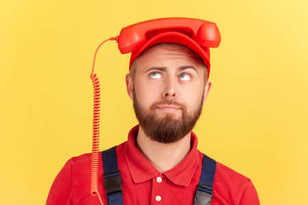 Portrait of funny worker man holding handset on his head and looking up, tired of calling for order service, wearing overalls and red cap. Indoor studio shot isolated on yellow background.の写真素材