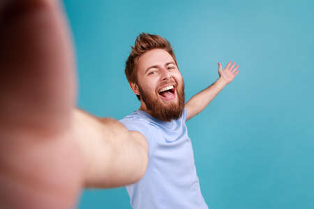 welcome. Portrait of excited satisfied man blogger POV, point of view of photo, laughing happily, and welcoming raised arm, expressing happiness. Indoor studio shot isolated on blue background.の写真素材
