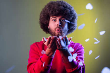 Portrait of hipster man with Afro hairstyle blowing heart shaped confetti, enjoying birthday or...