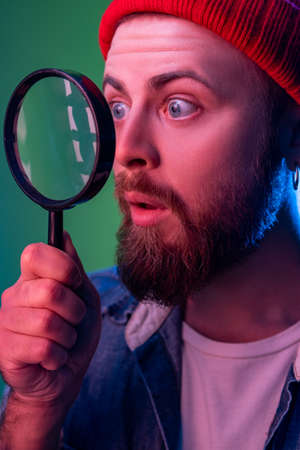 Side view of hipster man standing, holding magnifying glass and looking aside and having astonished shocked face. Indoor studio shot isolated on colorful neon light background.の写真素材
