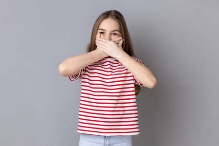 Portrait of dark haired little girl wearing striped T-shirt put hands on mouth, looking with fear in...