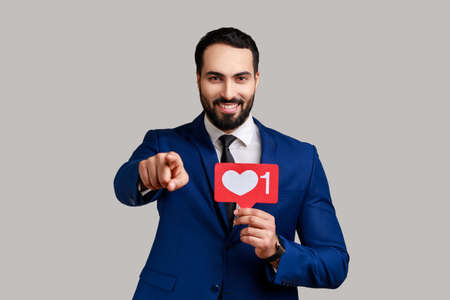 Satisfied man holding Heart Like icon of social media, notification button to follow, subscribe blog, pointing to camera, wearing official style suit. Indoor studio shot isolated on gray backgroundの写真素材