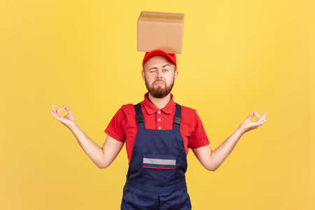 Portrait of relaxed courier man standing with cardboard box on his head, practicing yoga, looking at camera with one eye, calms down. Indoor studio shot isolated on yellow background.の写真素材