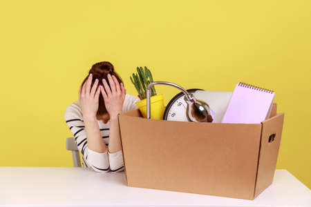 Sad upset desperate young woman office worker sitting at workplace with cardboard box with her...