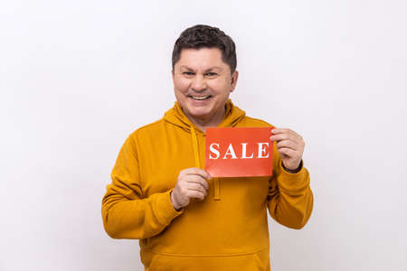 Portrait of delighted man holding sale card in hands looking at camera with toothy smile, black...