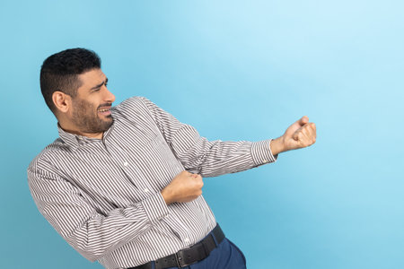 Side view of strong purposeful businessman with beard pulling invisible rope, showing his persistence and leadership qualities, wearing striped shirt. Indoor studio shot isolated on blue background.の写真素材