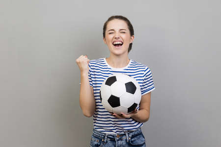 Portrait of woman wearing striped T-shirt screaming holding soccer ball, celebrating victory of favourite football team on championship. Indoor studio shot isolated on gray background.の写真素材