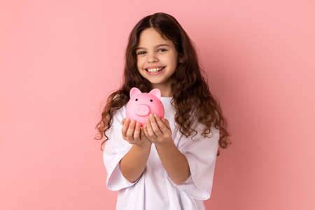 Portrait of little girl wearing white T-shirt holding pink piggy bank, looking at camera with...