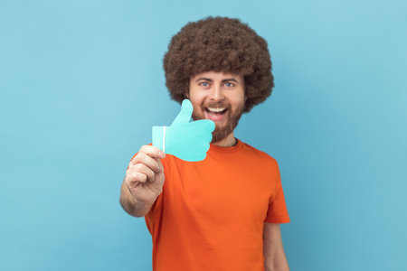 Man with Afro hairstyle wearing orange T-shirt showing thumbs up blue paper sign, liking and...