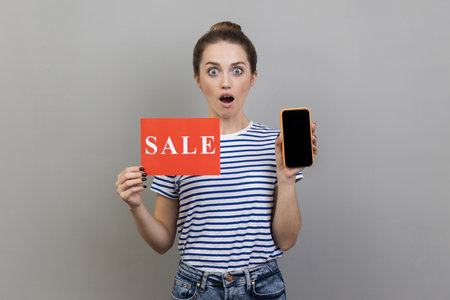 Portrait of shocked surprised woman wearing striped T-shirt standing with open mouth, holding sale card and cell phone with empty display. Indoor studio shot isolated on gray background.の写真素材