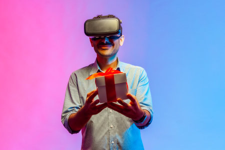 Man in VR virtual reality glasses holding wrapped gift box and smiling joyfully, giving present to camera, celebrating holiday. Indoor studio shot isolated on colorful neon light background.の写真素材