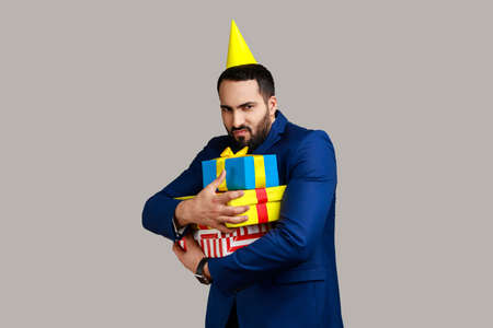 Sad bearded man with party cone on his head holding stack of presents in hands, being greedy to share gifts, wearing official style suit. Indoor studio shot isolated on gray background.の写真素材
