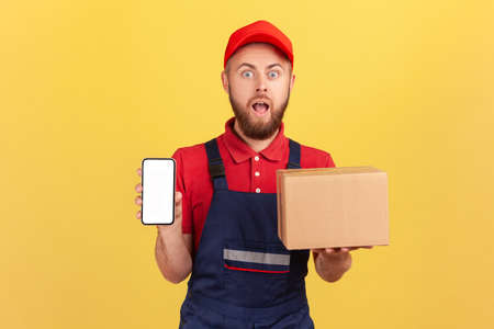 Portrait of surprised astonished courier wearing red T-shirt and blue overalls holding cardboard box and smart phone with empty screen. Indoor studio shot isolated on yellow background.の写真素材