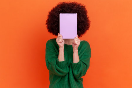 Portrait of unknown woman with Afro hairstyle wearing green casual style sweater standing holding organizer, hiding her face behind paper notebook. Indoor studio shot isolated on orange background.の写真素材
