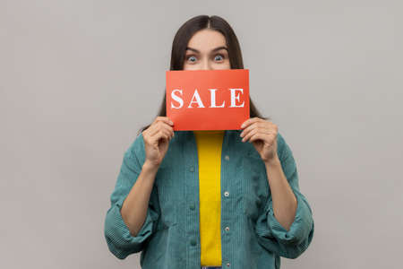 Portrait of young adult astonished surprised woman covering half of face with card with sale...