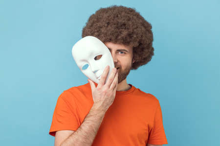 Portrait of man with Afro hairstyle wearing orange T-shirt peeping out white face mask, hiding his real feelings, pretending to be another person. Indoor studio shot isolated on blue background.の写真素材