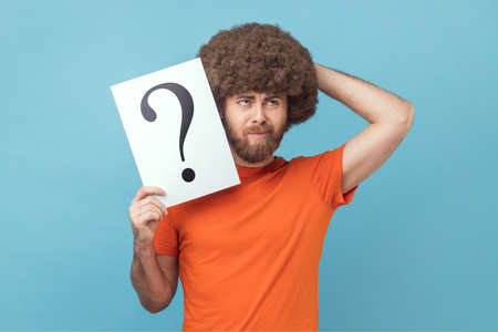 Portrait of pensive thoughtful man with Afro hairstyle wearing orange T-shirt holding question mark,...