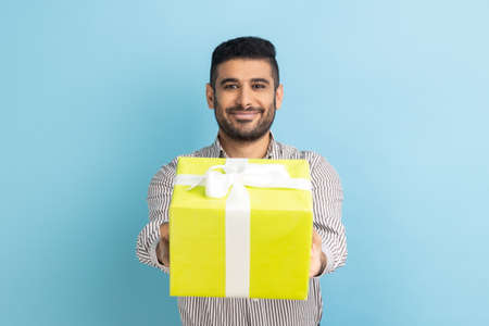 Portrait of smiling bearded businessman giving wrapped yellow gift box and smiling at camera, happy holiday, wearing striped shirt. Indoor studio shot isolated on blue background.の写真素材