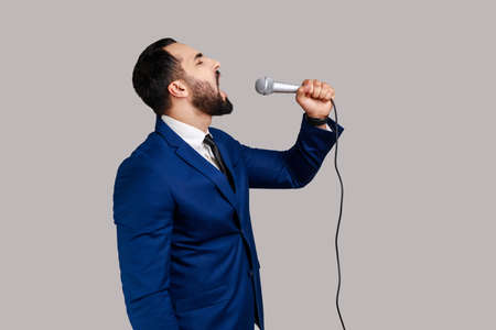 Side view of bearded man singing songs, holding microphone, singer making performance, keeps eyes closed, wearing official style suit. Indoor studio shot isolated on gray background.の写真素材
