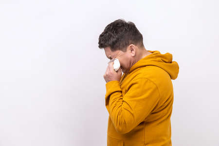 Side view of desperate middle aged man rubbing eyes, crying because of hopelessness and loneliness, nervous breakdown, wearing urban style hoodie. Indoor studio shot isolated on white background.の写真素材