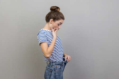 Portrait of beautiful surprised woman wearing striped T-shirt looking at her slim waist in big trousers with open mouth, successful weight loss. Indoor studio shot isolated on gray background.の写真素材