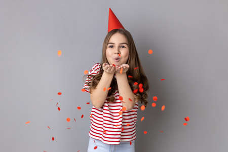 Portrait of happy little girl wearing striped T-shirt and party cone blowing heart shaped confetti,...