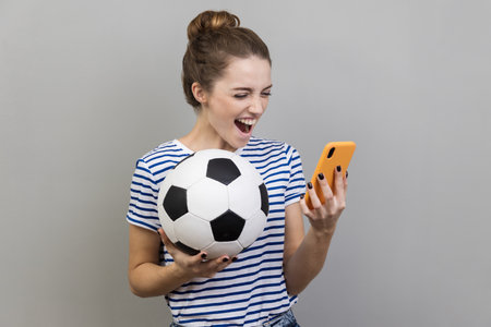 Portrait of excited positive happy woman wearing striped T-shirt holding soccer ball and smart...