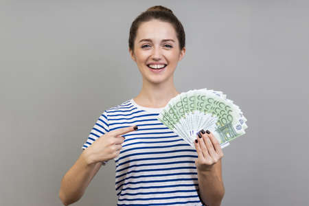 Portrait of woman wearing striped T-shirt pointing finger at fan of euros in her hand looking at...
