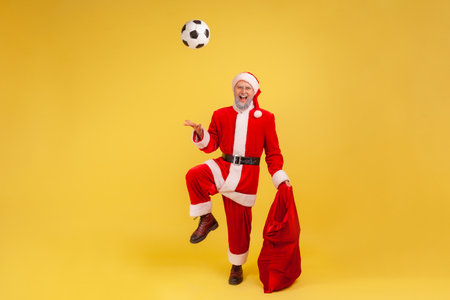 Full length of elderly man with gray beard wearing santa claus costume playing with soccer ball, holding red bag bags with new year presents. Indoor studio shot isolated on yellow background.の写真素材