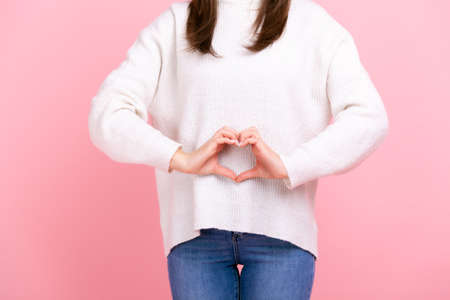 Unknown girl making heart shape in front of her belly as symbol of women health care, healthy eating, wearing white casual style sweater and jeans. Indoor studio shot isolated on pink background.の写真素材