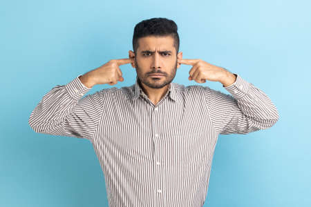 I dont want to hear this. Annoyed frustrated businessman closing ears with fingers ignoring information, irritating noise, wearing striped shirt. Indoor studio shot isolated on blue background.の写真素材