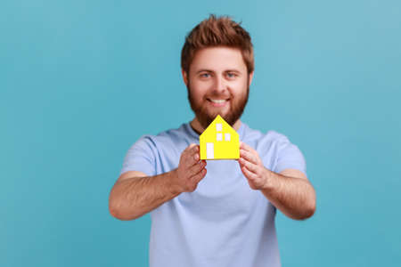 Portrait of happy bearded man holding in hands little yellow paper house looking at camera with pleasant smile, dreaming about own house. Indoor studio shot isolated on blue background.の写真素材