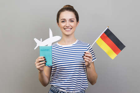 Portrait of happy satisfied woman with bun hairstyle wearing striped T-shirt standing with German flag and passport with paper plane, traveling. Indoor studio shot isolated on gray background.の写真素材